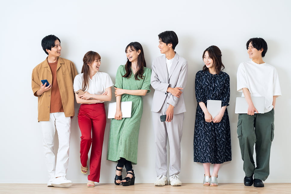 カジュアルな服装の若者6人が白い壁の前で笑顔で並んで立っている様子。全員がノートパソコンやスマートフォンを持ち、会話を楽しんでいるように見える